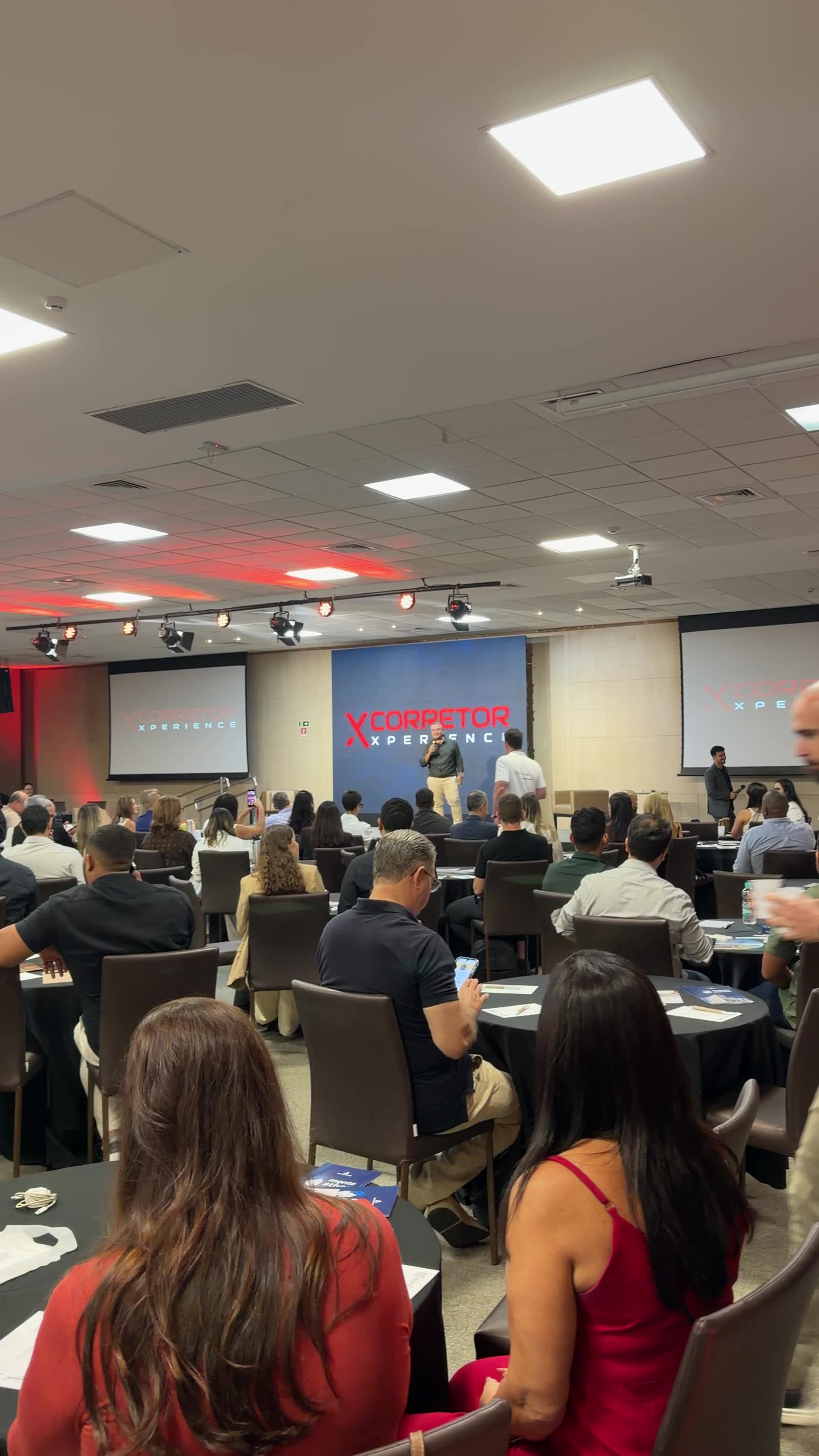 Salão do evento visto de trás da plateia: palco ao centro e telas laterais exibindo a marca Corretor Xperience.